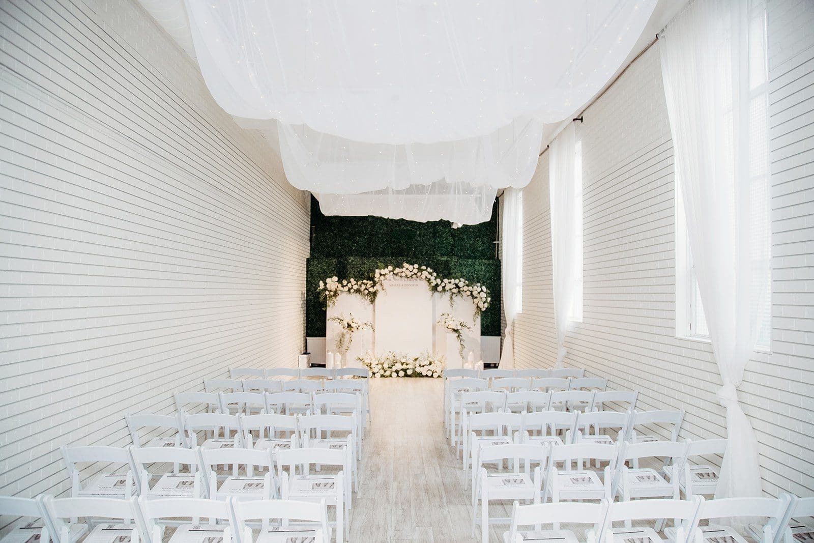 The main hall at Gather Walnut Creek set for a ceremony, white chairs down a center aisle facing a floral arch against the greenery wall